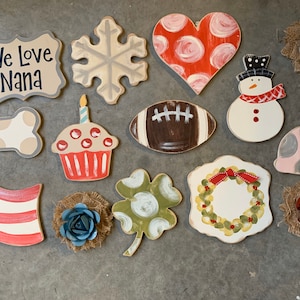 May include: A collection of 14 wooden painted shapes, including a heart, snowflake, cupcake, football, snowman, dog bone, shamrock, wreath, American flag, and a sign that says "We Love Nana".