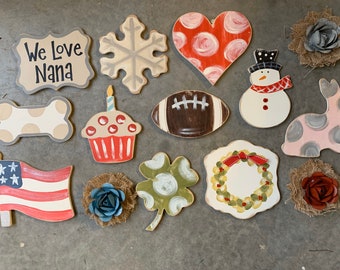 May include: A collection of 14 wooden painted shapes, including a heart, snowflake, cupcake, football, snowman, dog bone, shamrock, wreath, American flag, and a sign that says "We Love Nana".