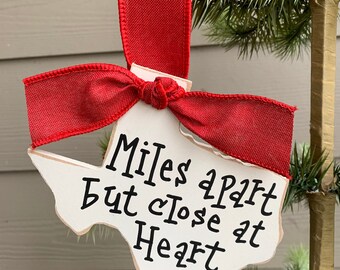 May include: White wooden ornament shaped like the state of Texas with the text "Miles Apart But Close At Heart" written in black. The ornament is tied with a red ribbon.