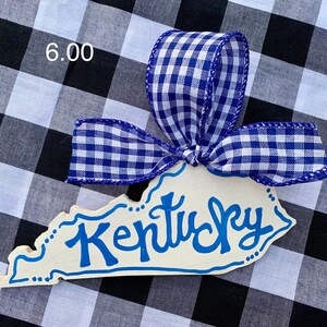 May include: A wooden ornament shaped like the state of Kentucky with the word "Kentucky" written in blue paint. The ornament is decorated with a blue and white gingham ribbon.