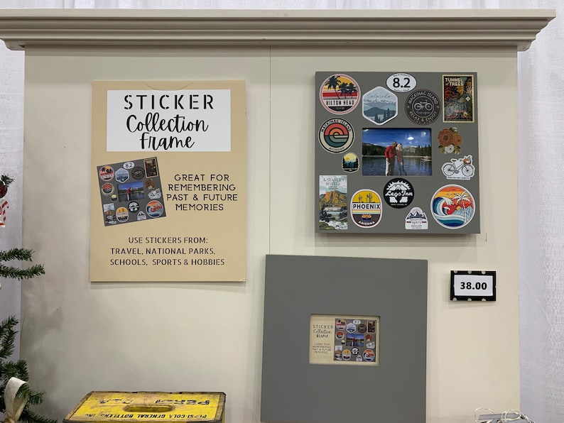 May include: A gray frame with a white background and a sticker collection inside. The frame is labeled "Sticker Collection Frame" and has a price tag of $38.00. The frame is designed to hold stickers from travel, national parks, schools, sports, and hobbies.