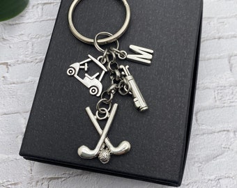 May include: A silver golf-themed keychain with a golf cart, golf clubs, a golf ball, and a golf bag charm. The keychain is attached to a silver key ring and has the letter "M" attached.