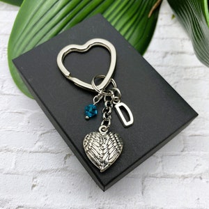 May include: Silver heart-shaped keychain with a charm of angel wings, a blue crystal, and a letter 'D'. The keychain is displayed on a black box.