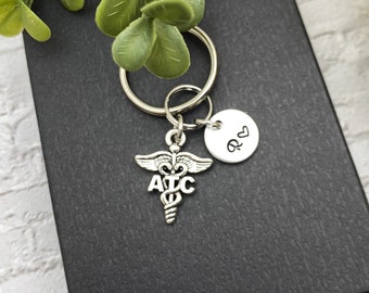 Può includere: Un portachiavi in metallo argentato con un ciondolo a forma di caduceo con le lettere ATC incise sul davanti. Attaccato all'anello c'è un piccolo disco rotondo in metallo argentato con la lettera R e un simbolo a cuore.