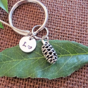 May include: Silver keychain with a small pine cone charm and a round charm with the letter "L" and a heart. The keychain is on a green leaf.