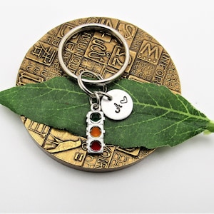 May include: A silver keychain with a miniature traffic light charm, displaying green, amber, and red lights. The keychain also features a small, round, silver charm with the initials "A" and "V" and a heart symbol. The keychain is displayed on a textured, bronze-colored surface with a green leaf.
