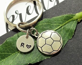 May include: A silver-toned metal keychain featuring a circular charm with the letter R and a heart symbol, alongside a round charm embossed with a soccer ball pattern. The charms are attached to a standard split ring.