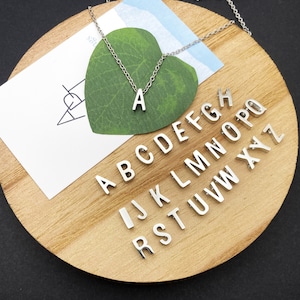 May include: A silver necklace with a letter A charm. The necklace is displayed on a wooden surface with a green leaf and a white card with the letters of the alphabet arranged in a circle.