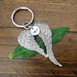 May include: Silver angel wing keychain with a heart-shaped charm engraved with the letter "M" and a heart. The keychain is on a green leaf.