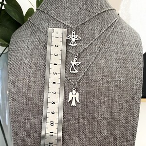 K&ouml;nnte beinhalten: Drei silberne Engelsanh&auml;nger-Halsketten auf einem grauen Stoffhintergrund. Die Ketten sind &uuml;bereinander gelegt und haben unterschiedliche L&auml;ngen. Die Anh&auml;nger sind alle im gleichen Design, aber in verschiedenen Gr&ouml;&szlig;en. Ein Lineal ist im Hintergrund sichtbar.