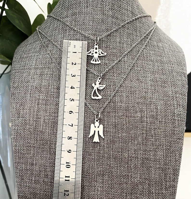 K&ouml;nnte beinhalten: Drei silberne Engelsanh&auml;nger-Halsketten auf einem grauen Stoffhintergrund. Die Ketten sind &uuml;bereinander gelegt und haben unterschiedliche L&auml;ngen. Die Anh&auml;nger sind alle im gleichen Design, aber in verschiedenen Gr&ouml;&szlig;en. Ein Lineal ist im Hintergrund sichtbar.