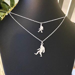 May include: A layered silver-toned necklace featuring two delicate chains, each adorned with a small, flat, cutout pendant shaped like a walking Bigfoot silhouette. The pendants are positioned at different lengths along the metallic link chains.