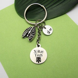 May include: Silver keychain with a round charm that says "X-Ray Tech" with a small ribcage image. The charm is attached to a silver chain with a small silver charm with a heart on it and a silver charm with a ribcage image.