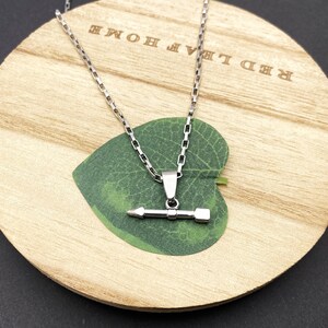 May include: A silver-toned metal necklace featuring a pendant shaped like a horizontal arrow or pencil. The pendant hangs from a delicate rectangular link chain. The jewelry piece is displayed against a green leaf-shaped cutout on a round wooden base.