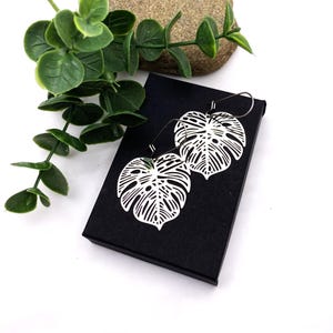 May include: A pair of silver-colored, leaf-shaped earrings with intricate cut-out details, displayed on a black box. The earrings are designed to resemble monstera leaves. Green foliage and a stone are in the background.