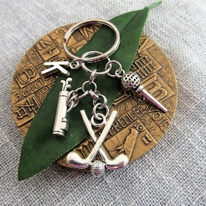 May include: Silver golf-themed keychain with a golf club, golf ball, and a microphone charm. The keychain is attached to a round, gold-colored metal tag with text.
