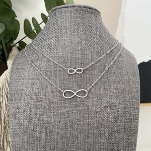May include: Two silver-colored infinity symbol necklaces displayed on a gray mannequin. The necklaces are of different lengths, with the larger one at the bottom and the smaller one above it. The infinity symbols are simple and elegant.