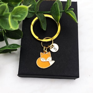 May include: A gold-colored keychain with a fox charm and a small, round charm with the letter "S". The fox charm is orange and white. The keychain is on a black box, with green leaves in the background.
