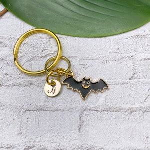 May include: A gold-toned metal keychain featuring a black enamel bat charm with a smiling face and a small circular gold-toned charm engraved with the letter N in a cursive script, attached to a circular split ring.