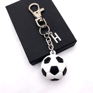 May include: A soccer ball keychain with a silver-tone clasp, chain, and the letter "H". The soccer ball is white with black pentagon shapes. The keychain rests on a black box.