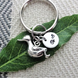 May include: A silver-toned metal keychain featuring a duck-shaped charm and a small circular disc charm. The circular disc is stamped with the letter Q and a heart symbol. The keychain is attached to a standard circular split ring.
