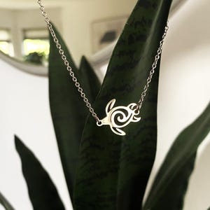 May include: A silver-colored necklace featuring a turtle pendant. The turtle design is a stylized outline with a spiral pattern on its shell. The necklace chain is delicate and hangs against a backdrop of green leaves.
