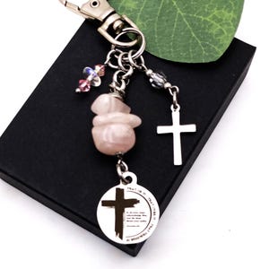 May include: A silver keychain with a lobster clasp, featuring a pink stone, clear and pink beads, and a silver cross charm. A round pendant with a cross and text is also attached. The keychain is on a black box.