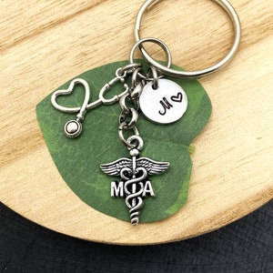 May include: A silver-toned metal keychain featuring a circular ring, a stethoscope charm with a heart-shaped earpiece, a round disc stamped with the letter M and a heart symbol, and a caduceus charm with the letters M and A integrated into the design.