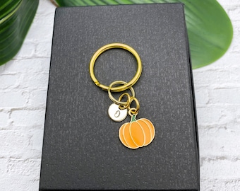 May include: A gold-toned metal keychain featuring a circular ring, a small round charm engraved with the letter O, and an orange enamel pumpkin charm with a green stem. The keychain is displayed on a black rectangular box.