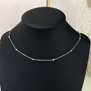 May include: A delicate silver-toned chain necklace featuring small, evenly spaced spherical beads along the length of the chain. The necklace is displayed on a black neck form, highlighting the metallic finish and the simple, repeating bead pattern.