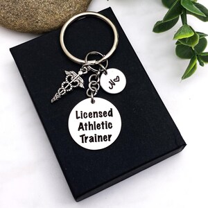 May include: A silver keychain with three charms. One charm is a caduceus symbol, another is a small round charm with the letter "M" and a heart, and the third charm reads "Licensed Athletic Trainer". The keychain is on a black box.