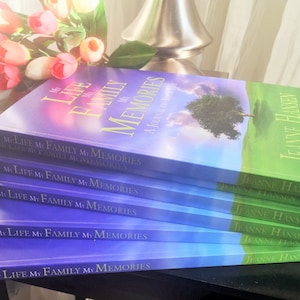 May include: A stack of six purple and blue books with the title "My Life My Family My Memories" and the author "Jeanne Hansen" on the cover. The books are stacked on a dark surface with a bouquet of pink flowers in the background.