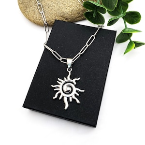May include: Silver-toned necklace featuring a sun pendant with a spiral center and radiating rays. The pendant hangs from a chain with elongated links. The necklace rests on a black box, with a stone and greenery in the background.