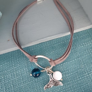 Puede incluir: Una pulsera de ante gris con un cierre plateado y un dije decorativo. El dije incluye una cola de ballena plateada, una cuenta azul y una cuenta blanca. La pulsera se muestra sobre una superficie azul texturizada.