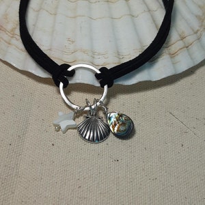Op de afbeelding: Zwarte choker ketting met een zilveren ring en drie bedels: een witte ster, een zilveren zeeschelp en een abalone schelp. De ketting wordt gepresenteerd op een grote witte zeeschelp.