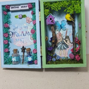 May include: A fairy-themed shadow box with a light blue and green frame. The box features miniature bottles, a fairy, a bird, and the text "Let your DREAM soar". The box is decorated with pink flowers and green moss, creating a magical scene.