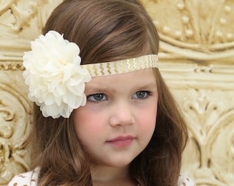 May include: A young girl with long brown hair wearing a white flower headband with a gold chevron pattern. She is wearing a brown and white polka dot shirt.