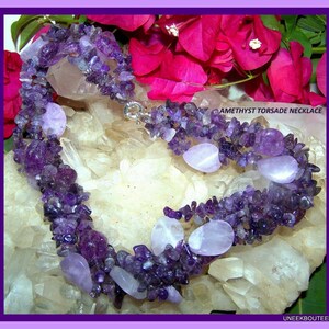 May include: A multi-strand amethyst necklace with a silver clasp. The necklace is made of small, irregular amethyst chips and larger, smooth amethyst stones. The necklace is displayed on a bed of white crystals.