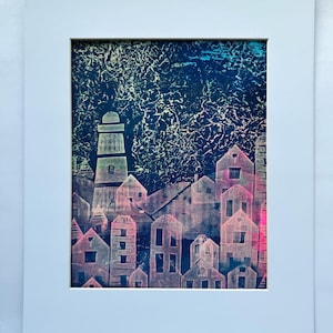 May include: Framed art print of a coastal town with a lighthouse and buildings. The artwork uses pink, blue, and white against a dark blue background. The print is matted in a white frame, suitable for home decor.