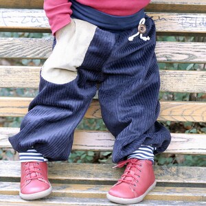 May include: A child wearing blue corduroy pants with a beige patch on the left leg and red shoes. The pants have a white drawstring and a brown button.