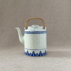 May include: White ceramic teapot with blue floral and geometric patterns. Features a woven handle and a spout. The teapot has a cylindrical body with small, evenly spaced holes. The base has a blue leaf design.