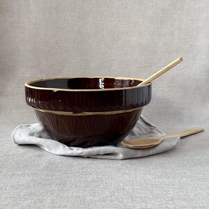 Peut inclure: Un grand bol mélangeur en céramique brun avec un motif décoratif autour du bord. Le bol est posé sur un tissu gris clair avec deux cuillères en bois à côté.