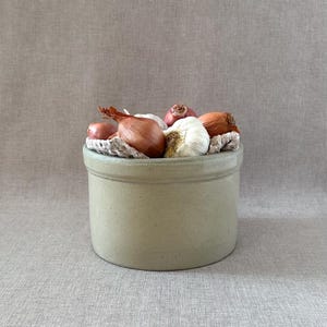 May include: A grey stoneware crock filled with shallots and garlic. The crock is sitting on a light brown linen fabric.