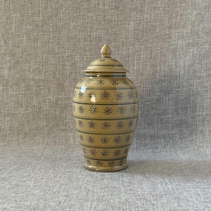 May include: A beige ceramic jar with a lid. The jar is decorated with horizontal blue stripes and floral patterns. The jar has a rounded body and a small, rounded knob on the lid.