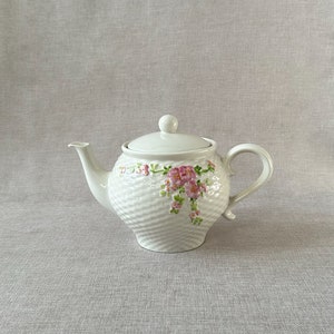 May include: White ceramic teapot with a floral design. The teapot has a textured surface and a lid with a knob. The flowers are pink and green.