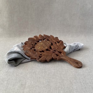 May include: A wooden trivet with a carved leaf design and a handle. The trivet is brown with gold accents and is lying on a gray fabric surface.