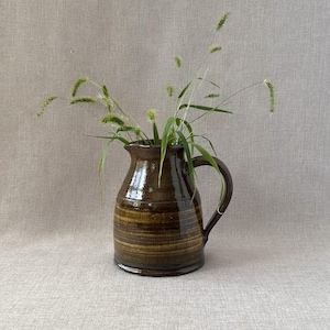 May include: A brown ceramic jug with a wide mouth and a handle. The jug has a striped pattern in a darker brown colour. The jug is filled with green grasses.