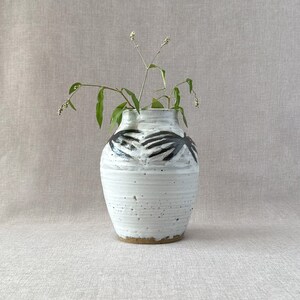 May include: White ceramic vase with black abstract design, featuring a rounded body and a wide opening. The vase is filled with green leafy stems and small white flowers.
