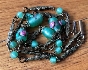 May include: A green and gold necklace with a chain and beads. The beads are made of glass and have a frosted finish. The necklace has a clasp closure.