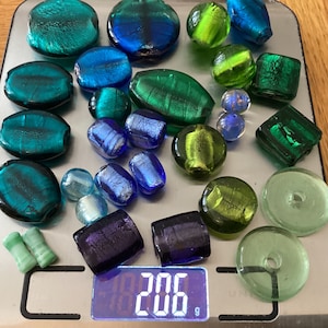 May include: A collection of green, blue, and purple glass beads with silver accents. The beads are on a digital scale that reads 206 grams.